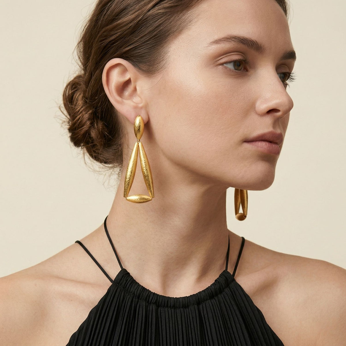 A woman with light skin and a tidy low bun frames bold Zeppelin Ivy Gold Clipon Earrings—gold-plated, hand-assembled in our West Hollywood studio. Sculpted geometry punctuates a black pleated top, echoing small-batch, eco-conscious artistry. Style Note: Refines both tailored suiting and evening silks.
