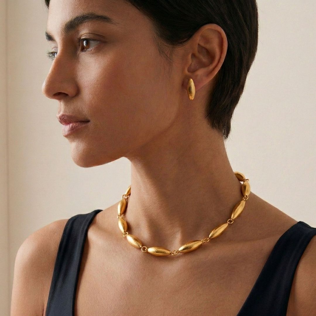 The Zeppelin Phoebe Gold Necklace by JAREDJAMIN Jewelry Online features eight elongated, textured oval links and small round rings, finished in matte gold with a toggle clasp and rectangular tag for vintage flair.