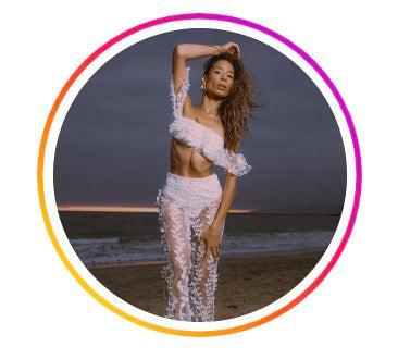 Woman posing on a beach, wearing a semi-transparent white outfit with floral details. She has long, curly hair and one arm raised, touching her head. The background shows a dark, cloudy sky and a strip of orange light on the horizon, framed within a colorful circular border.