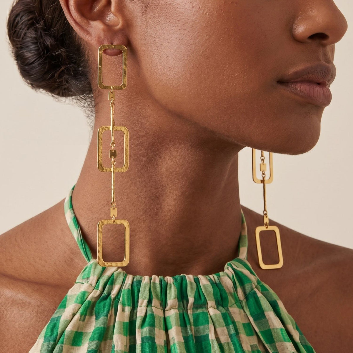 Side view portrait of a model wearing the 5-inch Solene gold rectangular drop earring paired with a green checkered top, demonstrating its shoulder-grazing length and light-diffusing hammered texture.