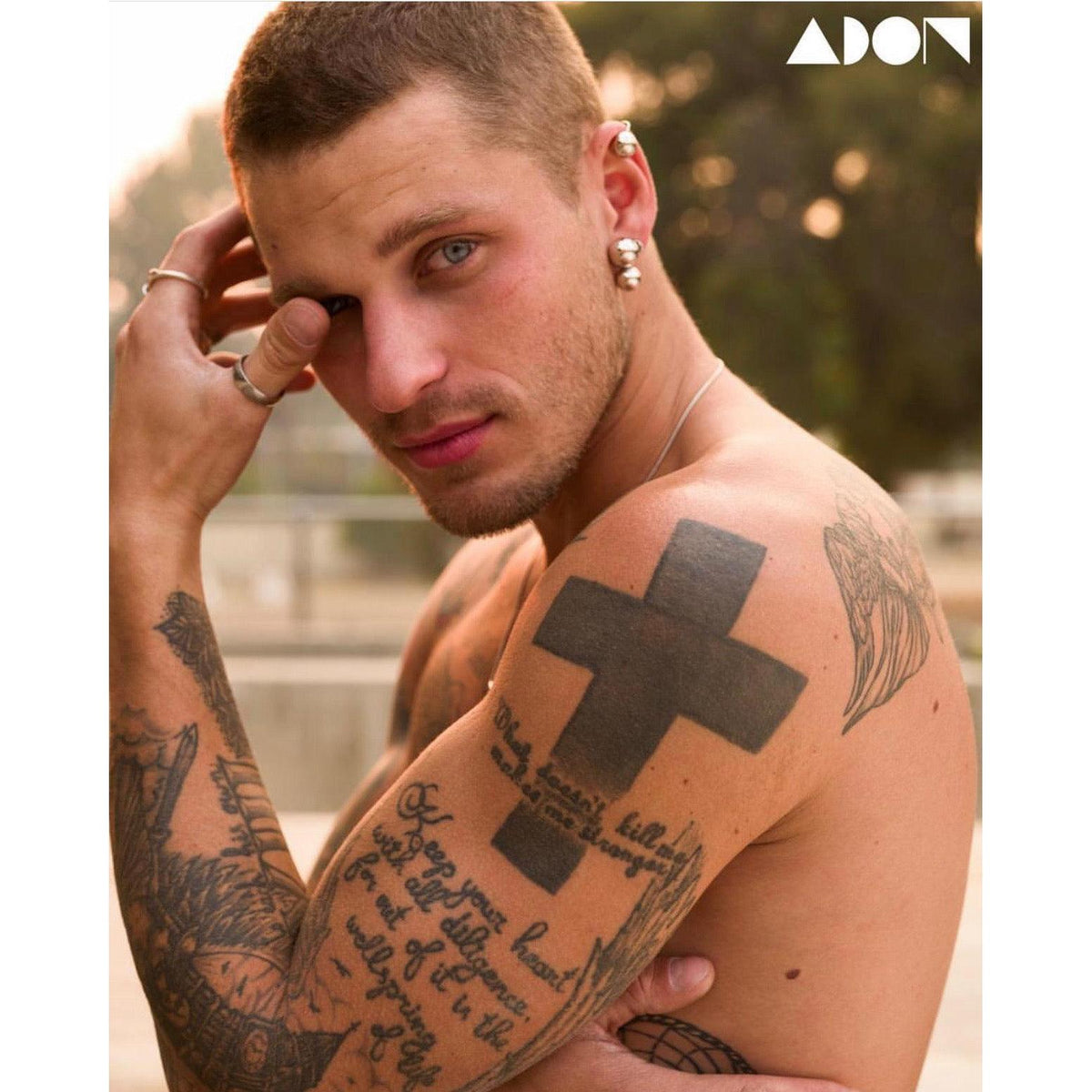 Un shirtless man con short hair y multiple tattoos looks into the camera, touching his face. He wears the JAREDJAMIN Joyería Online plata Orbita Ear puño pendiente. Los ADON logo appears at top right, con a softly blurred outdoor background.