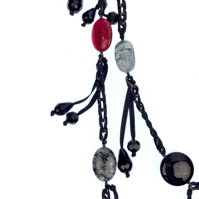 A close-up of the See You Saloon Necklace by JAREDJAMIN Jewelry Online shows large black, grey, and red quartz beads in oval and round shapes on braided black cord and gunmetal-tone chain with dangling small black beads, against a white background.